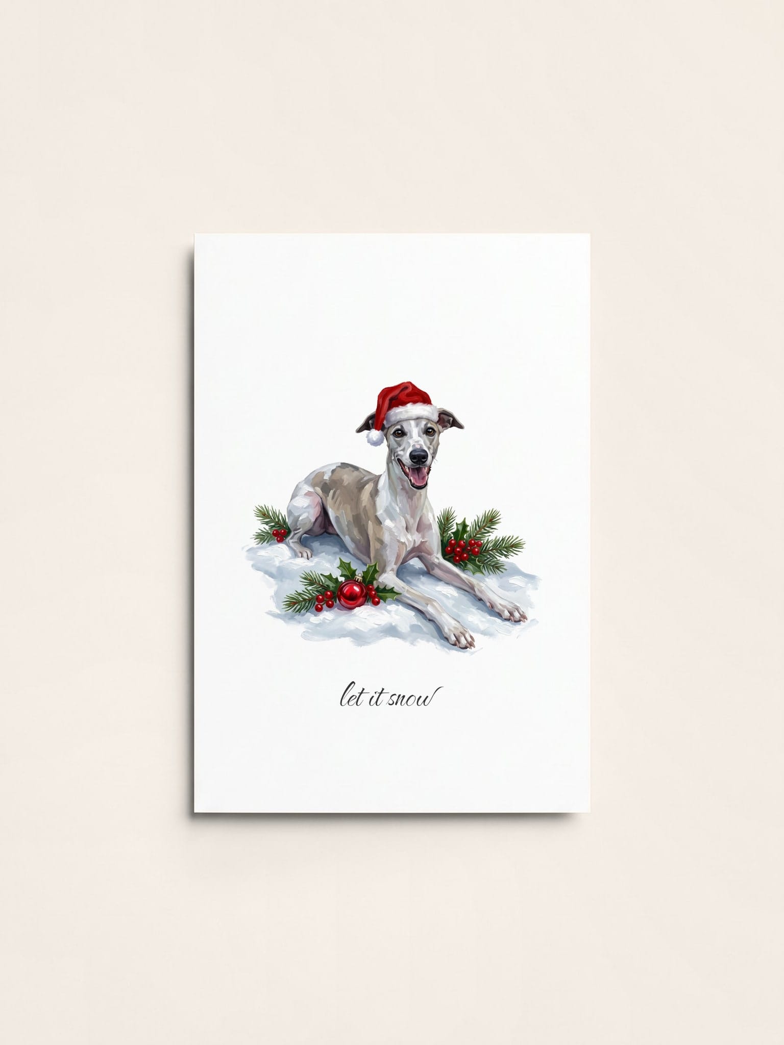 A Christmas card of a cream whippet in a Santa hat lying in the snow with holly and a red bauble